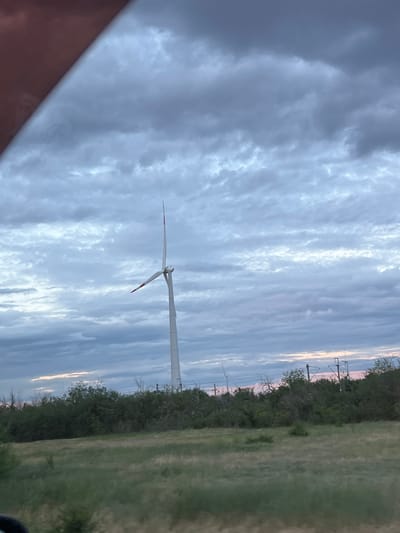 A single wind turbine stands tall against a cloudy sky. The turbine is white with red accents on its blades. The landscape below is a mix of green fields and dark green bushes. In the background, there are utility poles and wires. The image appears to have been taken from a moving vehicle, as indicated by the blurred foreground elements and the angle of the shot. The lighting suggests it may be dusk or dawn. There is no information available to determine the specific location in Zimovniki, Russia.