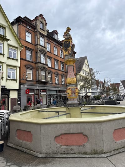 The image shows a partially obscured view of Biberach an der Riß in Germany, prominently featuring a stone fountain in the foreground. The fountain's structure is weathered with visible cracks, and details like its red-painted rectangular insets. Numerous chairs and tables are stacked around the fountain.

Behind the fountain are buildings, constructed of brick and various light-colored materials, displaying a mix of architectural styles, including windows with white frames. Above the buildings, the sky is overcast with a gray cloudy texture. There is a figure on scaffolding, suggesting maintenance or renovation work is in progress. Signage for Vodafone is visible on one building. The objects visible are tables and chairs in the plaza.