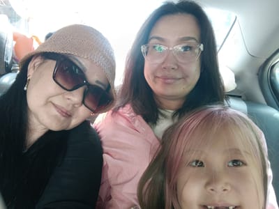 A selfie taken inside a vehicle shows three females. On the left, a woman wearing a straw hat and sunglasses smiles at the camera. Her dark hair is visible, and she is wearing a black top and a pearl earring. In the center, a woman with dark hair and glasses also smiles, wearing a pink collared shirt. On the right, a young girl with pink-tinted hair looks directly at the camera, her mouth open. The interior of the car is visible, including seats and a window. There is a bright light source behind the subjects, suggesting it is daytime. The image is likely taken in Yahşihan, Turkey, given the prompt's context, but no specific landmarks or environmental cues in the image confirm this location.