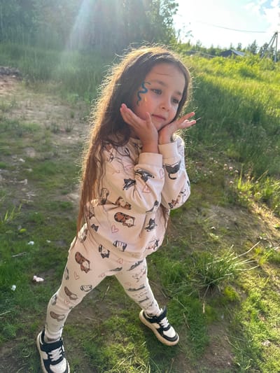 The image shows a young girl with long, dark hair adorned with blue face paint and glitter. She is wearing a light-colored sweatshirt and leggings with a pattern of cartoon animals, likely cats and tigers, and pink hearts. Her hands are placed near her face in a playful pose. She is wearing black and white sneakers with laces.

The setting appears to be outdoors in a grassy area, with a dirt path visible. The lighting suggests it is daytime, likely late afternoon, due to the warm glow and lens flare from the sun. The girl is looking towards the camera with a serene expression.

The background is out of focus but shows green grass and some vegetation. There are some small pink flowers scattered in the grass. In the upper left corner, a blurry electrical pole and wires are visible, hinting at a rural or suburban environment. The overall mood of the image is cheerful and playful, capturing a moment of a child enjoying the outdoors.

There is no visible text in the image. The location context provided is Oseyevo, Russia.