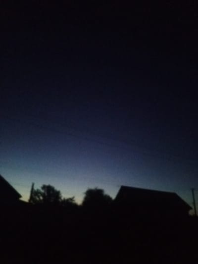 The image captures a silhouetted landscape against a dark, twilight sky. In the foreground, the dark outlines of several buildings and trees are visible. One building on the right has a prominent gabled roof. The sky transitions from a deep, dark blue at the top to a lighter, almost teal hue near the horizon. Faint horizontal lines, possibly power lines, are discernible against the darker parts of the sky. The scene appears to be set in a rural or suburban area, given the presence of houses and trees. The location is identified as Buraly, Russia, suggesting a view of dwellings and natural elements within that locale during dusk or dawn.