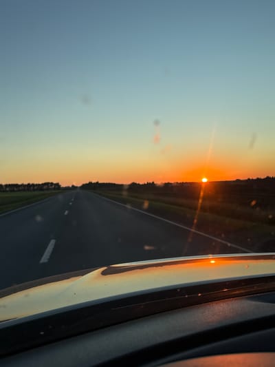 The image shows a view from inside a vehicle looking out at a highway during sunset. The sun, a bright orange orb, is setting on the horizon, casting a warm glow across the sky and reflecting on the vehicle's dashboard. The sky transitions from a pale blue at the top to a vibrant orange and yellow near the horizon. A line of trees forms a silhouette against the sunset on either side of the highway. The road ahead is paved with white dashed lines indicating lanes. The foreground shows the interior of the vehicle, with the reflective dashboard in focus. The scene suggests a drive through a rural or semi-rural area as day turns into night. The location is identified as the city of Морозовка, Russia.