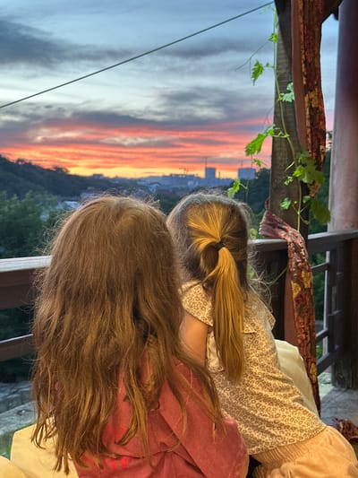 This image, taken in Myskhako, Russia, features two individuals, viewed from behind, observing a vibrant sunset. The sky is awash with streaks of orange, pink, and grey clouds, suggesting a dramatic twilight. Below the sky, a city skyline is dimly visible, nestled amongst dark green, tree-covered hills. The scene appears to be set on a balcony or porch, indicated by the wooden railing in the foreground and the draped fabric, possibly a tapestry, hanging overhead.
One individual has blonde hair tied back in a ponytail with a dark elastic band. They are wearing a light-colored top with a subtle, speckled pattern. The other individual, positioned in front, has long, flowing brown hair. They are wearing a reddish-pink garment, possibly a sweatshirt. The overall mood is one of quiet contemplation and appreciation for the natural beauty of the sunset. The time of day is clearly dusk.