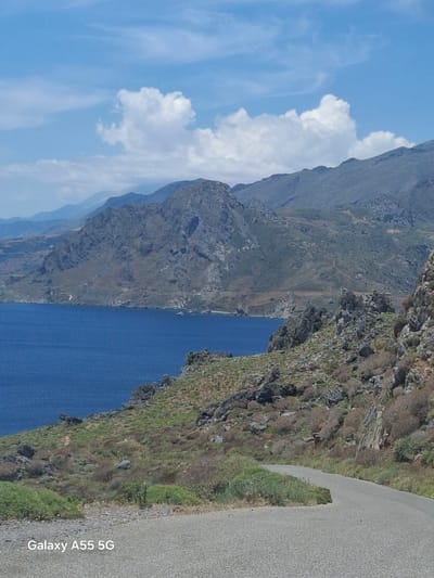 The image depicts a scenic view in Plakias, Greece, from an elevated perspective. The view captures a body of deep blue water, possibly a bay or inlet, nestled between rocky, mountainous terrain. The slopes are covered in sparse vegetation, with some areas exhibiting exposed rock formations. A road runs along the edge of the cliff on the right side of the image. The sky features a blue expanse with scattered white clouds. The camera used to take the image is identified in the corner as a "Galaxy A55 5G".