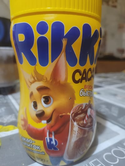 This is a close-up, eye-level, indoor shot of a yellow plastic container of "Rikki" brand cocoa-based drink. The container features a cartoon illustration of a character that appears to be a fennec fox wearing a red hooded sweatshirt with a blue "R" logo. The character is giving a thumbs-up gesture. The text on the container is in Russian and reads "Rikki Cacao" and "Вкусный быстрорастворимый" (Delicious instant). Below the character, there is text indicating "300 г" (300g). A glass of chocolate drink with a candy cane is depicted on the right side of the container. The container is placed on a surface that appears to be a table with a patterned tablecloth. The background is blurred and suggests an indoor setting, possibly a kitchen or dining area. The lighting is ambient and appears to come from above. This scene does not depict any specific location in the city of Hlusk, Belarus.