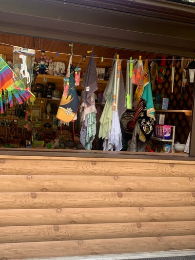 The image depicts an outdoor stall displaying various items for sale, potentially in the city of Arbuzov-Baran, Russia. The stall appears to be a wooden structure with shelves filled with merchandise. Prominently displayed are colorful textiles, plush toys, and assorted novelty items.

In the foreground, a wooden counter or facade with horizontal planks is visible. Behind this, a window or opening reveals the interior of the stall. Hanging from a line across the opening are several items, including a brightly colored kite, various patterned cloths or scarves, and what appears to be a toy gun in plastic packaging. Plush toys, including rabbits and other animals, are arranged on the shelves. Other items visible on the shelves include small boxes, bottles, and other trinkets. The background shows wooden shelving and walls. The overall impression is that of a small shop selling souvenirs or toys. There are no people visible in the image.