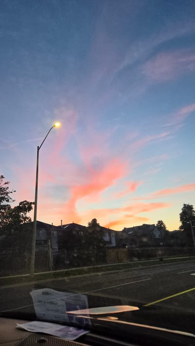 The image captures a sunset with vibrant pink and orange clouds against a gradient blue sky. In the foreground, a street lamp is illuminated, casting a yellow glow. Beyond the lamp, residential houses with dark roofs and chimneys are visible, partially obscured by trees. A street with white lane markings and a yellow line runs across the lower portion of the frame, indicating a road. The image appears to be taken from inside a vehicle, as evidenced by the reflection on a surface in the lower foreground, which shows what looks like documents or registration papers. The scene suggests a suburban or residential area in the city of Vaughan, Canada, during dusk.