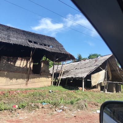 The image depicts a rural or semi-rural landscape in Payabeunyot, Indonesia, observed from a vehicle on a bright, sunny day. The sky is a clear blue with a few scattered white clouds, and multiple overhead power lines stretch across the upper frame.
The scene is dominated by two structures with traditional thatched roofs. The larger structure on the left features walls made from a combination of unrendered mud or clay and exposed red bricks. Its extensive thatched roof shows signs of wear and minor damage. Deep shadows are cast by the roof's framework onto the walls, indicating strong sunlight. An open window or doorway is visible in this structure.
To the right, a second, smaller structure with a sloping thatched roof is present, supported by wooden or bamboo poles. A light-colored piece of fabric or tarp hangs from its front. Within this structure, stacks of what appear to be unfired bricks or adobe blocks, alongside bundles of wood or sticks, are visible, suggesting a craft or building material production activity.
The foreground consists of reddish-brown dirt and patchy green grass, dotted with scattered litter such as plastic bags and wrappers. Lush green trees and foliage are visible in the background. Parts of the viewing vehicle are visible, including a dark interior section in the upper right and a rearview mirror in the lower right corner. The bright sunlight and clear sky indicate it is daytime. No visible text is present in the image.