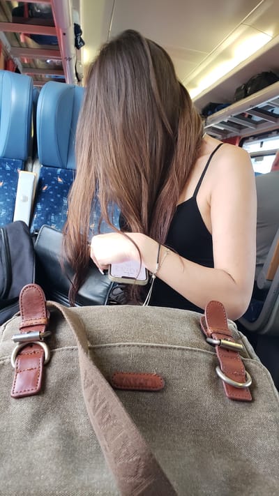 The image shows a woman with long, brown hair sitting in a train carriage. She is wearing a black tank top and is looking at her phone, which she is holding in her right hand. A brown canvas bag is in the foreground, with leather straps and buckles. The background shows blue train seats and luggage racks. The location is specified as Považská Bystrica, Slovakia.