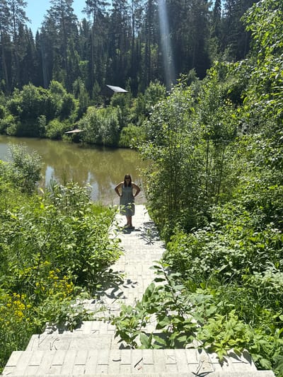 A woman stands on a tiled walkway leading down towards a lake. She is wearing a patterned dress and sunglasses, with her hands on her hips. The walkway is surrounded by lush green vegetation. In the background, a dense forest of tall trees borders the lake. The sky is clear and blue with a few sunbeams filtering through the trees. There is a small wooden structure visible in the distance amongst the trees on the opposite bank of the lake. The scene suggests a natural, serene outdoor setting, possibly a park or a recreational area near Makarovo, Russia.