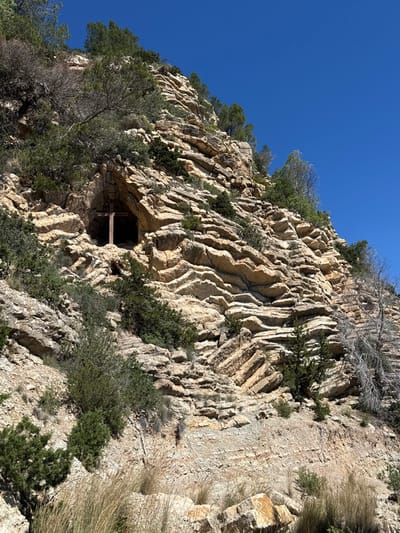 The scene depicts a steep, rocky hillside in Cala De Sant Vicent, Spain, under a clear blue sky. The terrain comprises stratified sedimentary rock, predominantly light brown and tan, exhibiting both horizontal layering and distinct chevron-like folding patterns. Sparse vegetation, including green evergreen trees and shrubs, is present on the upper slopes and within rock crevices; some desiccated vegetation and dry grasses are visible at the lower portions. A natural cave or deep recess is observed in the upper-mid left section of the rock face. A simple, weathered wooden cross stands upright within the entrance of this cave. No individuals or dynamic activity are present.
