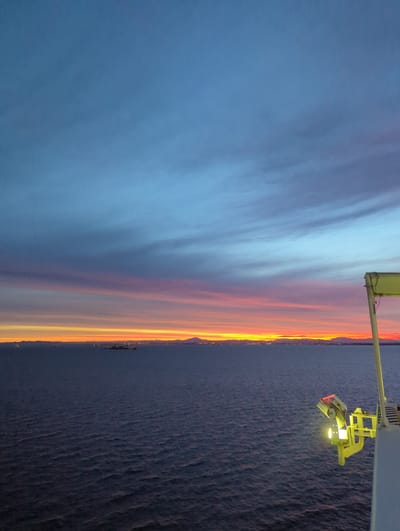The image depicts a sunset over the sea, likely viewed from a vessel. The sky displays a gradient of colors, transitioning from a deep blue at the top to streaks of pink, orange, and yellow near the horizon. A distant coastline with lights is visible, suggesting a city or populated area. Several ships and perhaps buoys are scattered across the water. In the foreground, a portion of a white structure with a yellow apparatus and a bright light is visible on the right side of the frame. This apparatus appears to be a spotlight or a similar lighting fixture. The sea is a dark, textured expanse, with small waves creating ripples on its surface. The scene conveys a sense of calm and twilight, with the artificial light in the foreground contrasting with the natural sunset. This image is likely taken from a ship in the waters off Yokkaichi, Japan, a major port city known for its industrial areas and coastal views.