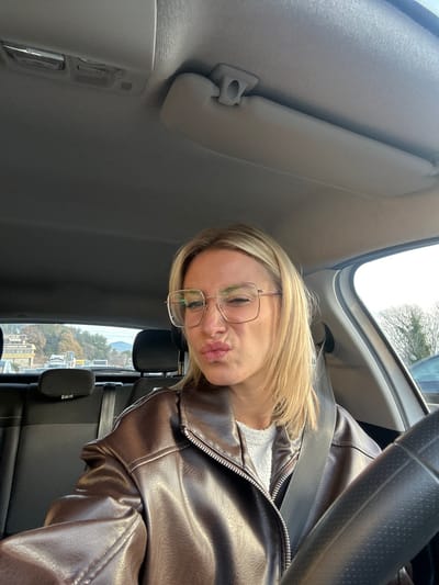 A blonde-haired female, wearing a dark brown zippered jacket over a light-colored top and large square-framed glasses, is seated in the driver's seat of a vehicle. She is taking a selfie, making a "duck face" expression with pursed lips and a slightly squinted right eye. A seatbelt diagonally crosses her chest. The car's interior features a light-colored headliner with an overhead light fixture and a sun visor, and dark-colored rear seats are visible behind her. A black steering wheel is partially visible on the right side of the frame. Through the rear window, a commercial building with a sign displaying "WORK" and partially visible text (likely "AT WORK AUTO") is visible. This business is situated at 505 Boulevard des Ferrayolles in Villeneuve-Loubet, France. A road or parking area in front of the building shows other vehicles, including a white truck. In the distant background, tree-covered hills or mountains are present under a bright sky. To the right of the vehicle, bare tree branches and bright sky are visible through a side window.