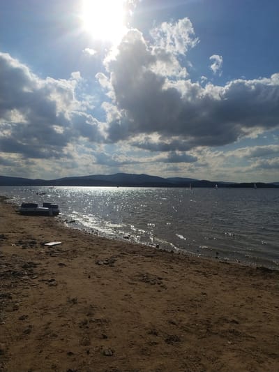 The image depicts a sunlit lake scene with a sandy shore. The sun is bright and high in the sky, casting a glare on the water and creating lens flare. Clouds are present in the sky, some are dark and appear to be rain clouds, while others are bright white and illuminated by the sun. The lake is calm with ripples on the surface, reflecting the sunlight. On the shore, there is an inflatable boat or raft pulled up onto the sand. In the distance, a line of hills or mountains can be seen under the clouds. Several sailboats are visible on the lake. The sandy shore in the foreground has tire tracks and scattered rocks and debris. The image does not show any people. The location is the Radslav Reservoir, near the village of Radslav in the Czech Republic.