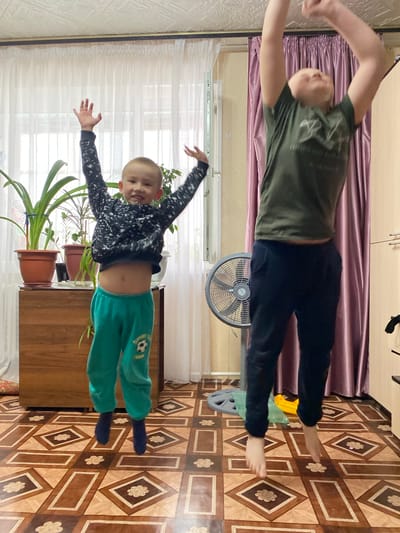 The image shows two boys in what appears to be an indoor setting. The older boy, wearing a green shirt and black pants, lies horizontally at the top of the image, his arms outstretched. The younger boy, dressed in a patterned top and green pants, is positioned horizontally at the bottom, with arms raised as well.
A window with white curtains is visible in the background. An electric fan stands behind the older boy. Several potted plants are also present. The flooring is a patterned brown and beige tile. The location is likely a home in Bely Klyuch, Russia, based on the request, but this cannot be confirmed with the image. The action portrayed is likely the boys pretending to fly or pose playfully.