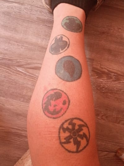 A leg is shown with five tattoos aligned horizontally. The tattoos are circular with varying designs and colors. The first is green with a dark interior, followed by a gray tattoo with a claw-like design inside. The next tattoo is blue with a dark interior. The fourth tattoo is red with a design resembling a dragon's head inside. The last tattoo is black with a sun-like design. The leg appears to be located on a wooden surface. The tattoos have no bearing on the location of Clarksville, Tennessee.