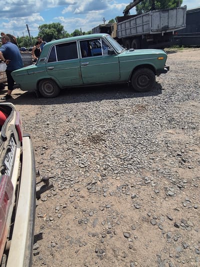 A green, four-door sedan, appearing to be a Lada VAZ-2101, is parked on a gravel lot. The car has visible rust and damage, particularly around the rear quarter panel. In the background, a large, utilitarian truck with a raised bed is visible, along with trees and utility poles. Three individuals are present, two men and a woman, near the left side of the sedan. One man in a blue t-shirt and black pants stands with his back to the camera. Another man, partially obscured, stands behind him. A woman with dark hair, wearing a black tank top and jeans, is also present. The scene appears to be outdoors in what could be a junkyard or a service area. The setting is consistent with the industrial outskirts of a city like Pidhaitsi, Ukraine, given the presence of such vehicles and the overall rugged environment.