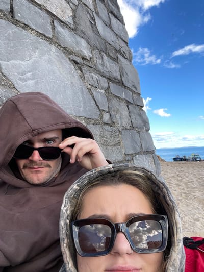 Two individuals, a male and a female, are depicted in a close-up, selfie-style photograph taken on a beach in Tenby, United Kingdom. They are positioned against a grey, multi-toned stone wall, which occupies the left side and top of the frame.
The male, positioned behind and to the upper left of the female, wears a plain brown hooded sweatshirt with the hood up. He has a dark mustache and is adjusting black sunglasses with his right hand, revealing his blue eyes which are directed towards the camera.
The female is in the foreground, partially cropped on the right and bottom. She wears a camouflage-patterned hooded sweatshirt with the hood up. Large, dark square-framed sunglasses cover her eyes, reflecting a blue sky with white clouds and a red-roofed building. A silver nose ring is visible on her left nostril. Blonde hair is visible beneath her hood.
To the right of the stone wall and behind the individuals, a sandy beach is visible. Further in the distance, the calm, blue sea is present under a bright blue sky with scattered white clouds. On the beach, near the waterline, two small fishing or utility boats are visible, one partially obscured, along with a yellow vehicle, possibly a small tractor. A small, dark object, potentially a distant bird or aircraft, is visible in the upper right sky. A red object, possibly a bag or garment, is partially visible in the lower right corner of the frame.