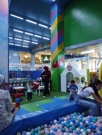 The image depicts an indoor play area for children, possibly in a shopping mall or dedicated entertainment center, located in Talas City, Kyrgyzstan. The environment is colorful and designed for play, featuring a ball pit filled with various colored balls, a small white playhouse with circular windows, and a bright blue padded boundary.
In the foreground, a young child is actively engaged in play, holding a large red cube. To the left, a woman wearing a floral patterned dress is seated, holding a yellow baby bottle. On the right side of the ball pit, a teenage girl in a white t-shirt and blue jeans is also seated on the edge.
Further back, several other children are visible. One child is near the playhouse, and another is running across the green and blue padded floor. An adult woman in a maroon hijab is standing and looking at her watch, appearing to supervise. Behind the play area, a wall with shelves filled with shoes and a blue shelving unit are visible, suggesting a waiting or shoe-storage area. The ceiling features a ribbed metal structure with numerous fluorescent lights, and a large, colorful striped pillar adds to the vibrant atmosphere. The overall scene suggests a lively and safe environment for children's recreation.