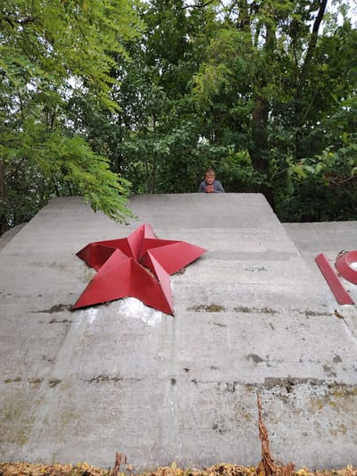 The image shows a close-up view of a concrete structure with a large red star ornament. A young boy is visible in the background, standing behind the concrete structure, peeking over the top. The structure itself appears to be part of an outdoor memorial or monument, indicated by the red star and what looks like parts of numbers on the concrete surface to the right. The environment is predominantly green, with dense trees and foliage surrounding the concrete element, suggesting it is situated in a natural or park-like setting. The concrete surface is weathered and shows signs of age, with some stains and cracks. The red star is geometrically shaped and prominently displayed on the concrete. The overall scene suggests a historical or commemorative site within the village of Balyko-Shchuchynka, Ukraine.