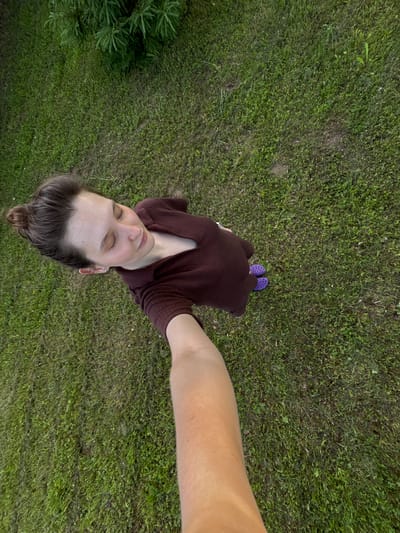 An overhead, high-angle selfie taken outdoors in Burdyata, Russia, shows a young woman standing on a grassy lawn. The woman, with her hair pulled up in a bun, is wearing a maroon sweater and purple Crocs. Her eyes are closed and her head is tilted back, suggesting relaxation or enjoyment of the sun. The background is a uniformly green lawn, with a few sparse plants visible in the upper left corner. The lighting suggests it is daytime, possibly late afternoon or early evening, given the soft, diffused light. The image appears to have been taken by the woman herself, extending her arm to capture the selfie.
