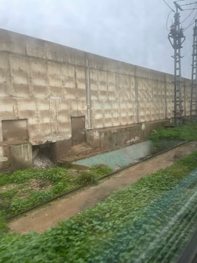 A tall, blocky concrete structure looms under a cloudy sky, with power lines visible at the top. Vegetation spills over an adjacent ditch. Several square openings punctuate the lower part of the wall. An overgrown drainage channel with blue tinges runs alongside a narrow path, densely lined with green plants. The angle suggests a ground-level, slightly tilted perspective.