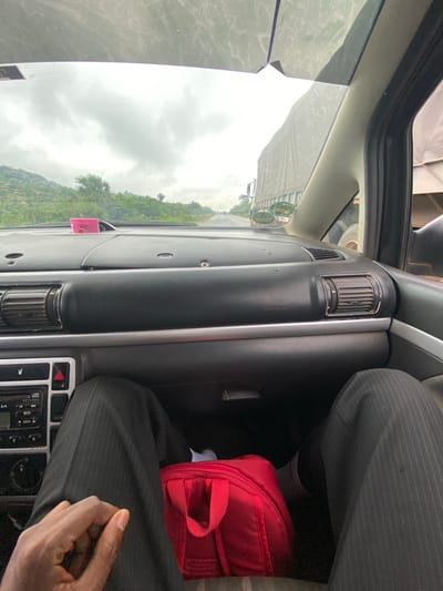 The image shows the interior of a car in Assob Hausa, Nigeria, with a view of a green hillside through the windshield. The dashboard is visible, along with air vents and a radio console. A red bag rests on the passenger's side. An individual wearing a pinstripe suit is seated in the car, visible from the waist down. Their hand is resting on their leg. The day is overcast.