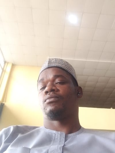 An adult male is depicted in a close-up, low-angle indoor photograph. The subject, of dark complexion, has a short beard on his chin and a dark ear stud in his left ear. He wears a light grey, long-sleeved garment with a collarless placket and a patterned light-colored cap. His gaze is directed forward, meeting the camera with a neutral expression. The background features a light-colored drop ceiling composed of square tiles, from which a single illuminated overhead light fixture emits a bright glow. The walls are light yellow or cream-colored, with a vertical structural element or window frame visible on the far left. The specific location within Kwi, Nigeria, cannot be determined from the visual information presented; the scene is consistent with an interior room.