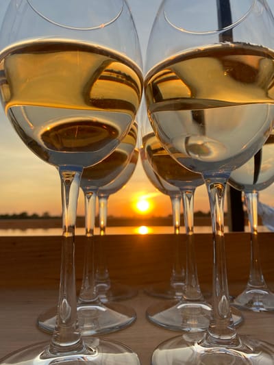 The image depicts multiple wine glasses filled with a golden liquid, arranged on a reflective surface. The background shows an out-of-focus sunset, suggesting an outdoor setting. The setting sun's reflection is prominent in the liquid within the glasses, creating a warm and luminous effect. The image does not contain information about any people, or any details indicating it was taken in Aramil, Russia.