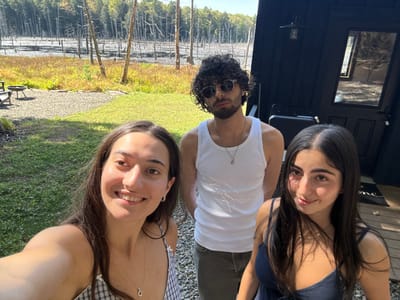 Three individuals are depicted outdoors on a sunny day in Otego, United States, in a rural or semi-rural setting. The scene features a dark-colored dwelling on the right and a marshy area with numerous bare tree trunks in the background.
From left to right:
1. A light-skinned female with long dark hair, smiling and looking directly into the camera. She wears a light-colored plaid camisole and a necklace with a circular pendant. She appears to be holding the camera, indicating a selfie.
2. A light-skinned male with dark curly hair, a beard, and sunglasses. He wears a white tank top and a necklace with a cross-like pendant. He stands facing the camera with a neutral expression.
3. A light-skinned female with long dark hair, looking slightly to her right with a subtle smile. She wears a dark blue camisole.
The foreground consists of green grass and a gravel pathway. To the far left, a metal fire pit and an outdoor chair are visible on the gravel. The dark dwelling on the right features a black exterior, a black door with a rectangular window, and an exterior light fixture mounted above the door. A "Welcome" mat is present at the doorstep. The background is dominated by a marsh or pond filled with numerous leafless, skeletal trees or stumps, beyond which a dense line of green-foliaged trees is visible under a clear, brightly lit sky.
