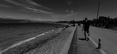 In a monochrome image, a person with a backpack walks on a paved road beside a pebble beach. The road runs parallel to the sea, which is to the left of the frame. The sea is dark and choppy, with a white wave breaking near the shore. The beach is covered in small, grey pebbles. In the distance, mountains are visible, softened by atmospheric haze. Several other people are walking further down the road, in the same direction as the main subject. Trees line the right side of the road, and a utility pole with a lamp is also present. The sky is bright, with wispy clouds. The image appears to be taken in a coastal area. The location is likely in the Municipal Unit of Aegio, Greece, given the terrain and vegetation.