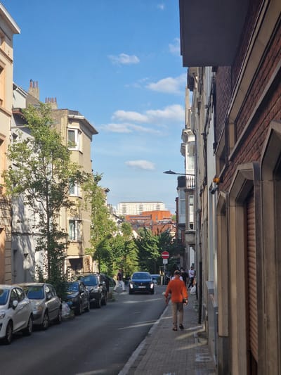 The image depicts a street scene in Saint-Josse-ten-Noode, Belgium, with buildings lining a paved road. The perspective is rotated, suggesting the photo was taken looking upwards. Several cars are parked along the street, and a person in an orange shirt is walking on the sidewalk, facing away from the camera. Another person in white is also visible further down the street. Trees with green foliage are present, and the sky is a clear blue with scattered white clouds. A traffic sign with a red circle and a diagonal bar is visible on the left side of the street. The overall impression is of a quiet, daytime street in a European city. The architecture of the buildings suggests an urban environment. The lighting indicates it is daytime, likely late morning or early afternoon, based on the shadows cast.
