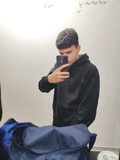 An indoor mirror selfie depicts a young male in Hrishteni, Bulgaria, positioned centrally within the frame. The male subject, likely a teenager or young adult, has short, dark brown hair with a frontal fringe. He is wearing a dark, possibly black, hooded sweatshirt that exhibits multiple small white or light-colored specks. His left hand holds a black smartphone, featuring a multi-lens camera module, positioned to capture his reflection. The subject's gaze is directed towards the smartphone, and his eyebrows are slightly furrowed. In the foreground, a dark blue duffel bag or gym bag, partially open, rests on a dark surface. A blue, possibly textured, fabric is visible inside the bag. The background consists of a plain, light-colored, textured wall. A white, decorative element, resembling cursive script, is mounted on the wall in the upper left quadrant. A small, light-colored circular container with blue accents is partially visible on the far right edge of the frame, near the bottom.