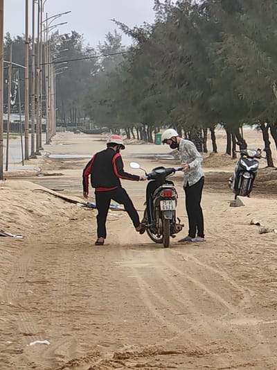 An outdoor scene in Thuận An Ward, Vietnam, depicts two males interacting with a motorcycle on a sandy path under an overcast sky. The individual on the left, wearing a black and red jacket, black pants, dark flip-flops, and a red and white helmet, is positioned with his left foot on the kickstart mechanism of a black motorcycle (license plate 75-H1 6929), appearing to attempt to start it. His right hand is near the motorcycle's left handlebar. The second individual, on the right, wears a patterned long-sleeved shirt, black pants, light-colored shoes, a white helmet, and a dark face mask, standing beside the motorcycle and holding a dark object in both hands. Behind them and to the right, another black motorcycle is parked upright on the sandy ground. The foreground and midground consist of sandy terrain with visible tire tracks and scattered debris. In the background, a paved road or promenade extends to the left, lined by a series of tall streetlights. A dense row of pine trees forms a green backdrop across the upper-midground. A green trash bin is visible under the trees, and a dark boat-like object is faintly discernible further down the paved area.