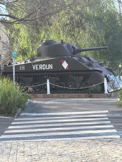 The image depicts an outdoor scene in La Valette-du-Var, France, featuring a military tank serving as a monument. The central subject is a dark olive drab or green tank, identifiable as an M4 Sherman variant, positioned on a raised platform. The tank's hull prominently displays the white word "VERDUN" and a diamond-shaped emblem featuring red, white, and blue segments, alongside a white 'C' marking. Its main gun barrel is aimed towards the right side of the frame.
The tank is partially encircled by a low barrier composed of white bollard-style posts connected by chains. To the right of the tank, several tall, rectangular, highly reflective panels are angled towards the viewer. Lush green foliage, including bushes and what appears to be dense ground cover, grows in front of and around the tank's base. In the background, deciduous trees and dense green vegetation are visible. A green informational sign with white text, partially legible as "A LA MEMOIRE D'ANDRE PRIET 192A-1950", is mounted on a pole to the left of the tank.
In the foreground, a pedestrian crosswalk with white stripes is painted across an asphalt road surface. The immediate foreground consists of a cobblestone pavement. No people, active subjects, or specific interactions are present in the scene. The lighting suggests daytime conditions with natural illumination.