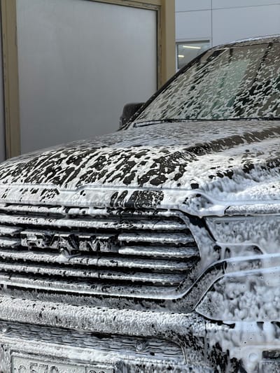 The image presents a close-up, low-angle perspective of a dark-colored vehicle, likely a Land Rover Defender, being thoroughly washed at what appears to be an indoor or covered car wash facility. The vehicle is seen rotated, appearing to stand vertically in the frame, coated almost entirely in a thick, white layer of cleaning foam.
The front grille, characterized by prominent vertical slats, is visible on the right side of the image, heavily caked with suds. Within the grille structure, partially obscured by the foam, the letters "DEFEN" are faintly discernible on a horizontal bar, identifying the vehicle as a Defender. The foam adheres to the vehicle's contours, creating a high-contrast pattern against the dark paintwork, with some areas showing intricate textures and drips.
To the left, a portion of the vehicle's side panel or window reflects the surrounding environment, showing streaky foam patterns over a darker, glossy surface. Reflections of interior lights or parts of the car wash structure can be seen on this reflective area.
The background consists of light grey or white walls, possibly tiled, indicating an indoor setting. A rectangular frame, possibly a window or a door, with a lighter interior, is visible in the lower left quadrant of the image. The scene captures the activity of a vehicle being pre-soaked or cleaned with foam in Bezborodovo, Russia.