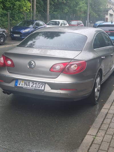 The image shows the rear of a grey Volkswagen Passat CC parked on a wet asphalt street. The car has a German license plate that reads "V NA 2107". In the background, other cars are visible, including a blue Ford Fiesta with a license plate "V SY 3003". The scene appears to be in a residential area with trees and buildings. The weather seems to be overcast and rainy, as indicated by the wet surfaces and water droplets on the cars.