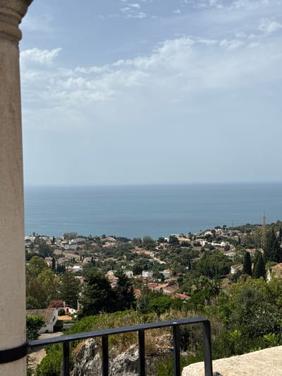 The image shows a panoramic view overlooking the coastal city of Benalmádena, Spain. In the foreground, a stone column and a black metal railing frame the scene. The middle ground is filled with a dense, green landscape of trees and vegetation, interspersed with numerous white-roofed houses and buildings with terracotta tiles, indicative of Mediterranean architecture. A winding road is visible snaking through the hillside. Beyond the cityscape, the vast expanse of the bright blue Mediterranean Sea stretches to the horizon under a hazy, pale blue sky with scattered white clouds. The lighting suggests it is daytime, likely late morning or early afternoon. No people or animals are visible in the image.