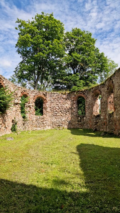 The image shows the ruins of a stone building with arched windows. The walls are made of rough-hewn stones and bricks. Green grass covers the ground inside and outside the ruins. Trees with lush green foliage are visible behind the walls, and a partly cloudy blue sky is in the background. The scene appears to be a historical site. This is a view of the Embūte Castle Mound ruins in Embūte, Latvia.