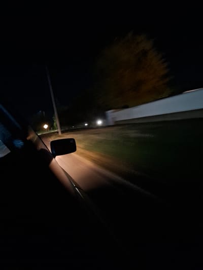The image depicts a scene at night, taken from inside a moving vehicle, likely on a road with a speed limit sign visible in the distance. The foreground is dominated by the dark interior of the car, with a portion of the windshield and a side mirror visible. The road ahead is illuminated by the car's headlights, creating a blur of motion. On the right side of the frame, a long, white, low-lying structure with a slight upward slope is visible, possibly a wall or a building. Beyond this structure, a dark, indistinct area suggests trees or vegetation. The overall impression is one of a nighttime drive through a semi-urban or rural area. There are no people visible in the image. No specific landmarks or details indicate the location within Yambol, Bulgaria.