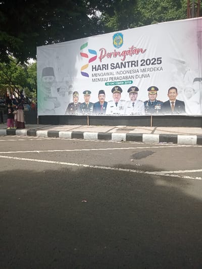 This image depicts an outdoor scene in Nganjuk, Indonesia, focused on a large rectangular white banner commemorating "HARI SANTRI 2025" (Santri Day 2025), scheduled for October 22, 2025. The banner, supported by a dark base, displays the slogan "MENGAWAL INDONESIA MERDEKA MENUJU PERADABAN DUNIA" (Guarding Independent Indonesia Towards World Civilization), a colorful abstract logo, a circular emblem, and faint background images. Seven portraits of adult males, identified as officials including the Acting Regent of Nganjuk (PJ. BUPATI NGANJUK), are prominently featured at the bottom. To the left of the banner, two adult females, both wearing headscarves, stand with two young children. Further left, a group of indistinct individuals is partially visible under dense green trees. The foreground consists of dark asphalt pavement with white painted lines and a black-and-white striped curb, containing minor litter.