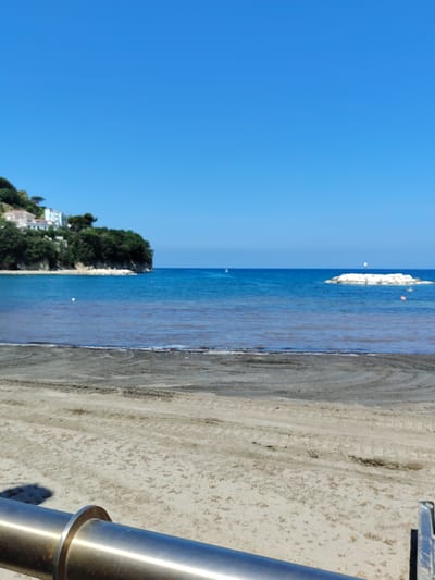 This media file captures a serene coastal scene in Agropoli, Italy. The image is dominated by a vast, clear blue sky, suggesting a bright, sunny day. Below the sky, the tranquil blue sea stretches towards the horizon.

On the left side of the frame, a lush green cliff juts out into the water, adorned with white buildings and dense trees, hinting at a picturesque seaside town. The shoreline in the foreground is a sandy beach, with tire tracks visible on the wet sand, possibly from a vehicle that recently traversed the area. A metallic railing or pipe is prominent in the lower part of the image, partially obscuring the sand and sea.

In the sea, a white barrier made of large rocks is visible, likely a breakwater, with a small flag or marker on top. Further out, there are hints of buoys and possibly a distant boat, suggesting the area is used for recreation or navigation. The water itself appears calm, with gentle waves lapping the shore.

The overall impression is one of peacefulness and natural beauty. The lighting suggests it is daytime, likely late morning or early afternoon, given the brightness and the clear blue sky. There is no visible text in the image. The scene evokes a sense of calm and relaxation, typical of a Mediterranean beach setting.