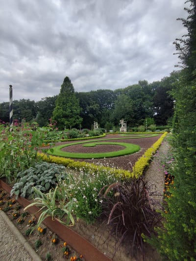 The image shows a formally landscaped garden with manicured hedges, flower beds, and a central circular design element. In the foreground, there are various plants including feathery, dark purple ornamental grass, pale green foliage plants, and small orange flowers. A rusty metal border separates the flower beds from a gravel path. In the mid-ground, a circular pathway with mown grass borders a darker, mulched area arranged in concentric circles. Two classical statues, one depicting a person on horseback, are visible in the distance within the garden. A tall, dark green conifer tree stands behind the statues, and a dense line of trees forms the background. The sky is overcast with gray clouds. A black and white striped pole with a red top is visible on the left side of the image. This garden is located at Anholter Schloss in Isselburg, Germany.