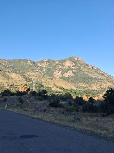 This image shows a rocky, mountainous landscape with sparse vegetation, under a clear blue sky. A paved road runs along the right side of the frame, disappearing into the distance. The sun appears to be setting or rising, casting long shadows and illuminating the mountainside with a warm, golden light, suggesting it is either early morning or late afternoon.
In the mid-ground, a few structures are visible, including what looks like a small building with a brown roof and a white electrical tower. There are also some animals grazing on the grassy slopes. The overall scene evokes a sense of peacefulness and solitude. The location is identified as Shatin, Armenia. There is no visible text in the image.