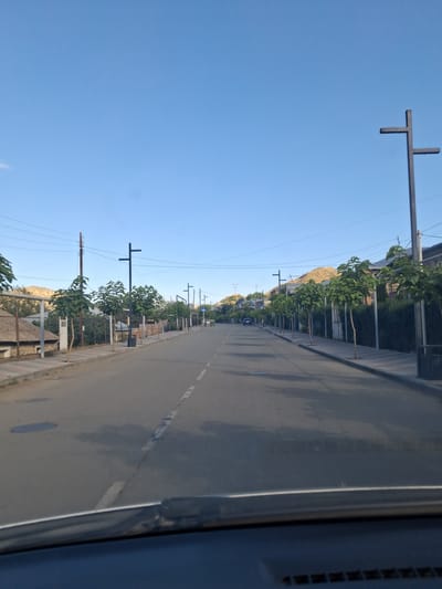 The image depicts a street scene in Vayk, Armenia, under a clear blue sky. The street is paved with asphalt and has a dashed white line marking the center. Along the right side of the road, there is a sidewalk bordered by a fence and lined with young trees. Modern streetlights with multiple arms are spaced along the sidewalk. Further back, there are buildings and dense vegetation. On the left side of the road, there is also a sidewalk, a fence, and some houses with thatched roofs. In the distance, hills with sparse vegetation are visible. A few cars are present on the road, and power lines run overhead. The overall scene suggests a residential or semi-urban area in Vayk.