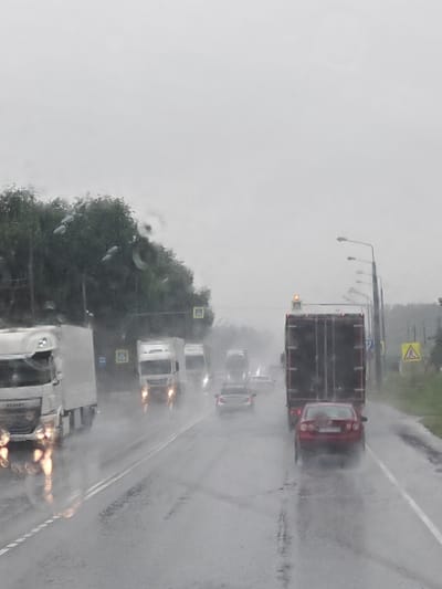 The image depicts a rainy day on a highway, likely near Gavrilovskoe, Russia, given the context. The scene is dominated by overcast skies and wet road conditions, with visible raindrops on the windshield. Multiple vehicles are present, including several large trucks and smaller passenger cars. The trucks are white and dark-colored, some with trailers. A red sedan is prominently featured in the middle of the frame, appearing to be in motion. Yellow triangular road signs with black symbols are visible, indicating potential hazards or warnings. Streetlights are also present along the roadside. The overall atmosphere is dim and wet, typical of inclement weather. There are no visible people in the image.