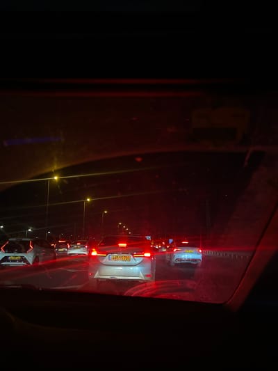 The image shows a traffic jam at night on a road in Sdot Dan Regional Council, Israel. The perspective is from inside a vehicle, looking forward through the windshield. Several cars are visible, with their headlights and taillights illuminated. Streetlights line the road, casting a yellowish glow. The license plates of some cars are visible, one reads "445.55.701" and another reads "657.03.402". The overall scene is dark, with reflections on the windshield obscuring some details.