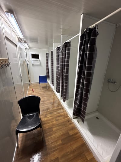 The image shows a row of shower stalls lined up along the right wall of a narrow room, located in the city of Mazkeret Batya, Israel, at the coordinates 31.833 latitude and 34.82 longitude. Each stall is enclosed with a white frame and black and white checkered shower curtain. The shower on the far right has a white shower pan and a chrome shower head with a hose. To the left of the showers are water-stained wooden floorboards and a black plastic chair. In the background, on the left side of the frame, is a white wall with a row of hooks. On the far left wall, there is a doorway that leads to a room with a white wall and a window. In the center of the room, near the shower stalls, is a blue plastic chair.