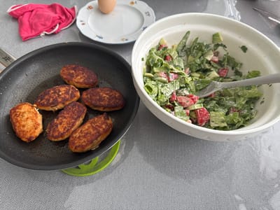 A close-up, overhead shot captures a meal being prepared and served. In a black non-stick frying pan, several oval-shaped, golden-brown cutlets are being cooked. They are arranged in a neat row, with a green silicone mat underneath the pan. To the right, a white ceramic bowl is filled with a fresh salad. The salad appears to be made of chopped lettuce, tomatoes, and possibly cucumbers, all coated in a creamy dressing. A gray fork rests in the salad bowl, suggesting it's ready to be served or is in the process of being eaten.

In the background, a small white plate with a decorative floral rim holds a single, hard-boiled egg. Next to it, a folded pink cloth with white trim lies on a textured gray tablecloth. The overall scene suggests a home-cooked meal, likely lunch or dinner, given the combination of a main dish (cutlets) and a fresh salad. The lighting indicates it's daytime. The image is sharp and well-lit, focusing on the food. There is no visible text in the image. The setting appears to be a domestic environment, possibly a kitchen or dining area.