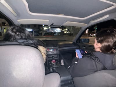 An interior view of a vehicle at night in Glyka Nera Municipal Unit, Greece, shows two occupants in the front seats. The driver, positioned on the left, is partially visible from the rear, exhibiting long dark hair and wearing a dark jacket with a fur-lined hood. The front passenger, on the right, is viewed from the back, displaying short dark hair and wearing a black padded jacket. This passenger is actively holding and observing two smartphones; the left device displays a light-themed user interface, and the right device shows a dark-themed interface. The vehicle's interior features a gray headliner, gray cloth seating, and a dark dashboard. The center console contains an automatic gear shifter with a small red decorative object attached. Through the vehicle's windows, an urban street environment is visible under night illumination, with discernible streetlights and building facades, one of which displays a partial blue sign with "BANK" text.